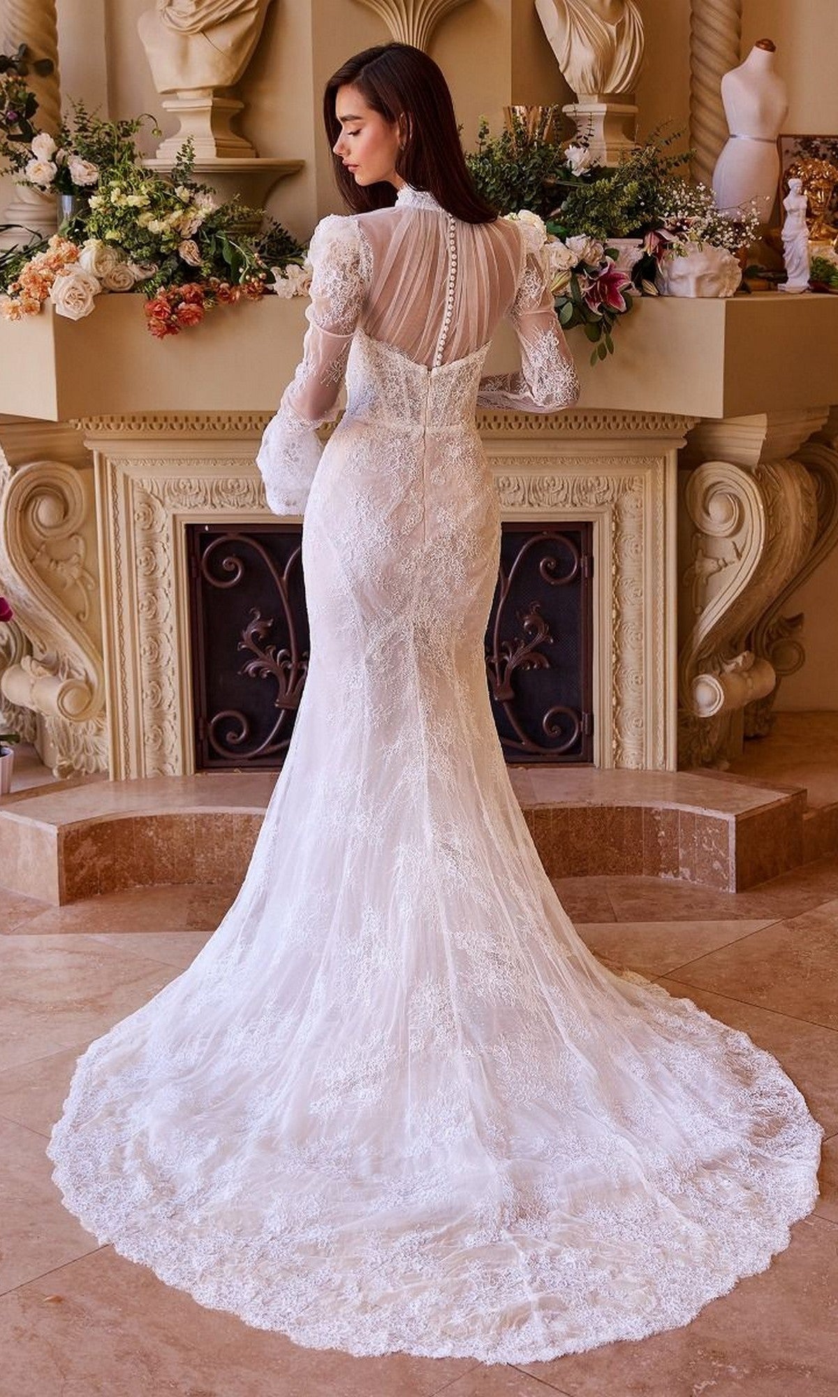A woman in the Andrea and Leo WL042 long wedding dress, featuring a fit-and-flare silhouette, lace long sleeves, and a train, stands facing away before an ornate flower-adorned fireplace.